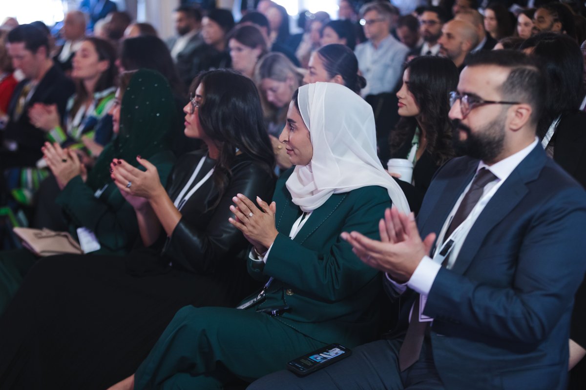 Rows of seated conference attendees applauding, with a woman in a white headscarf and green suit in the foreground