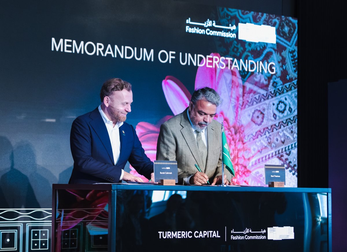 Two representatives signing documents onstage beneath a Memorandum of Understanding backdrop for Turmeric Capital and the Fashion Commission