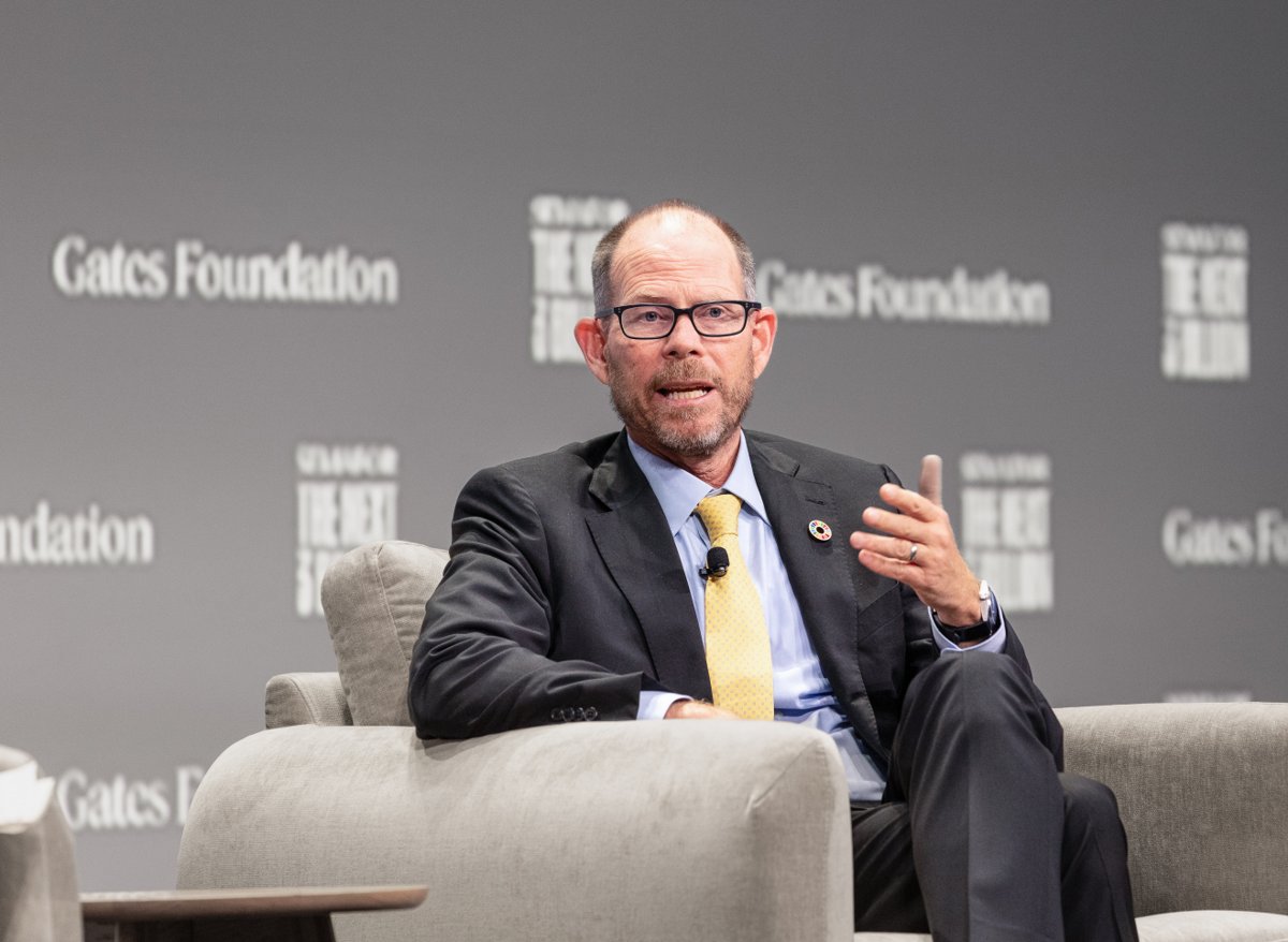 A seated speaker in a dark suit and yellow tie gesturing in front of a Gates Foundation backdrop