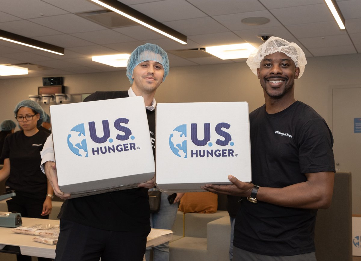Two volunteers wearing hairnets and holding U.S. Hunger boxes at a food-packing event