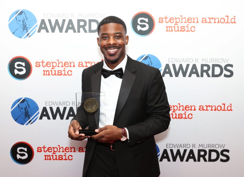 Smiling honoree in a tuxedo holding a Murrow Award in front of the event backdrop