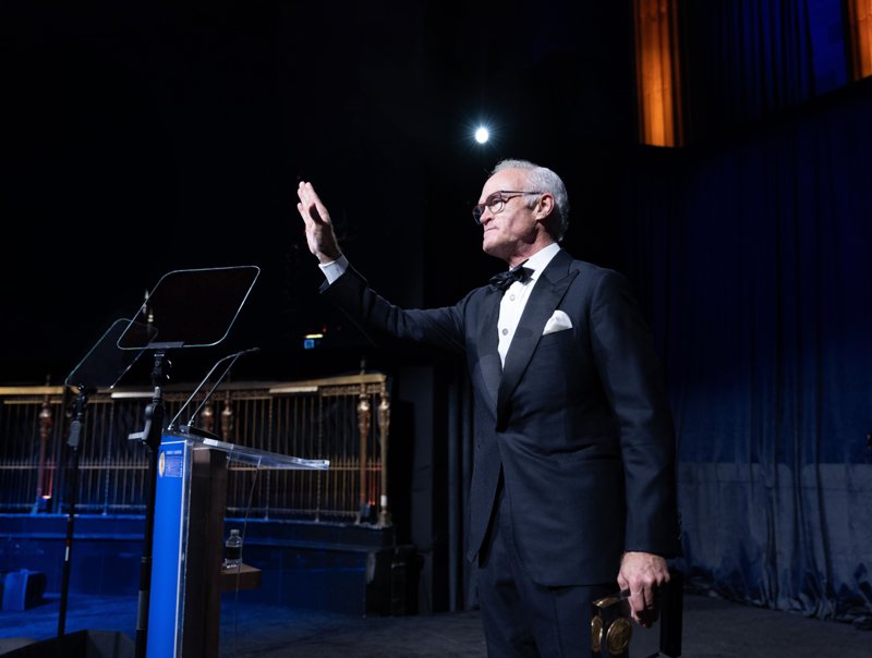 Gala speaker in a tuxedo raising his hand beside a lectern on stage