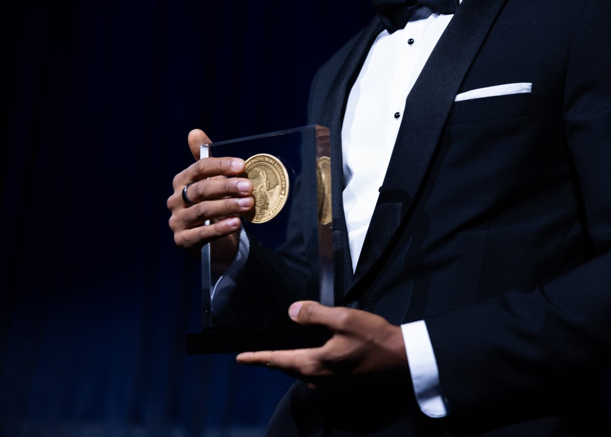 Close-up of a person in a black tuxedo holding a glass Edward R. Murrow Award with a gold medallion