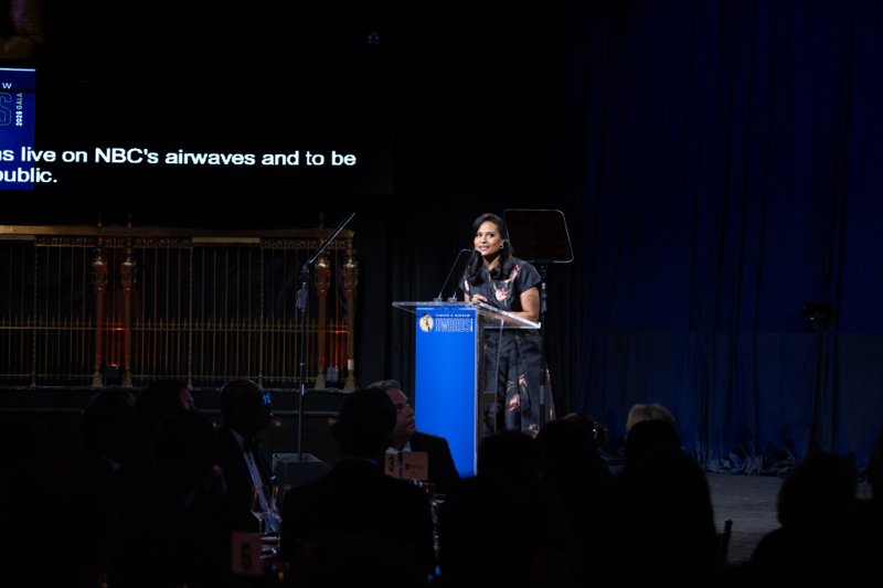 Woman addressing the gala audience from a blue lectern beneath stage lighting