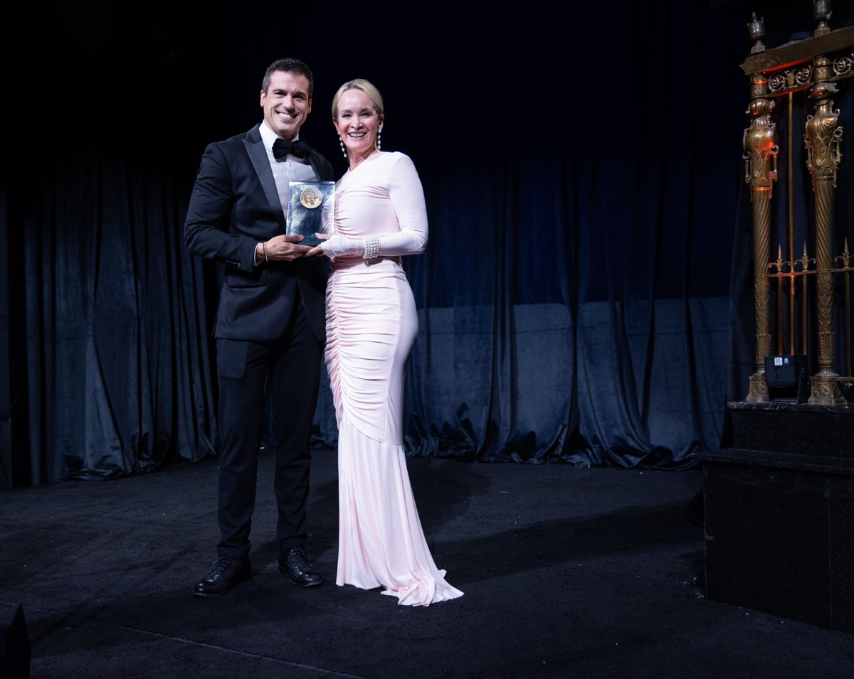 Man in a black tuxedo and woman in a pale pink gown smiling together on stage while holding an Edward R. Murrow Award